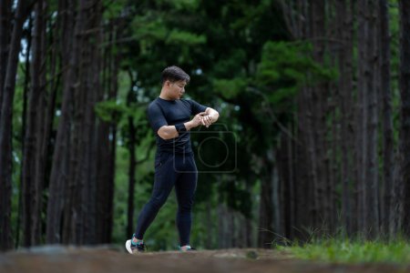 Téléchargez les photos : Chemin asiatique coureur court en plein air dans la forêt de pins route de terre pour l'exercice et l'entraînement des activités tout en vérifiant la montre intelligente sur la perte de calories atteindre un mode de vie sain et concept de remise en forme - en image libre de droit