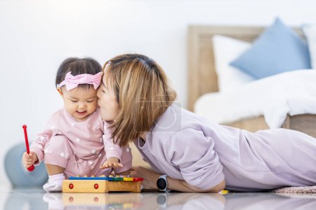 Téléchargez les photos : Mère asiatique kssing sa jolie fille bébé souriante avec bloc jouet en bois tout en passant du temps de qualité dans le lit pour le bonheur de la famille et la parentalité - en image libre de droit