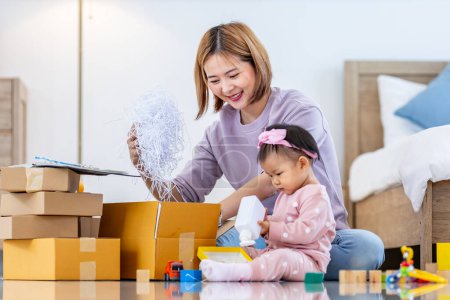 Photo for Young Asian mother is packing the merchandise product package while working at home ready to deliver while her toddler kid is playing around with her toy - Royalty Free Image