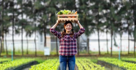 Foto de Mujer asiática agricultora está llevando la bandeja de madera llena de verduras orgánicas recién recogidas sobre su cabeza en el jardín para la temporada de cosecha y la alimentación saludable - Imagen libre de derechos