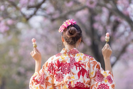 Foto de Mujer japonesa en vestido tradicional de kimono sosteniendo dulce postre de hanami dango mientras camina en el parque en el árbol de flor de cerezo durante el festival de sakura de primavera - Imagen libre de derechos