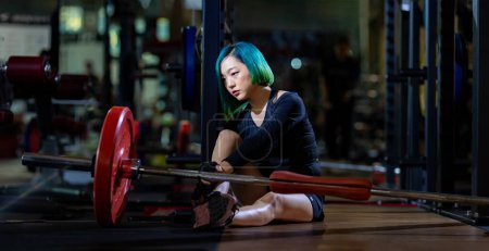 Foto de Mujer asiática se enfrenta al síndrome de sobreentrenamiento después del entrenamiento con pesas dentro del gimnasio con fondo oscuro para el ejercicio y el concepto de acondicionamiento físico - Imagen libre de derechos