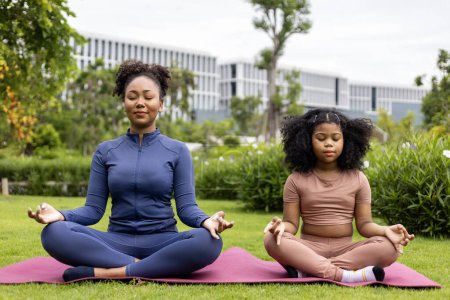 Foto de Mujer afroamericana y su hija en traje de yoga están practicando relajadamente ejercicio de meditación en el parque para alcanzar la felicidad de la paz interior sabiduría para la mente sana y el alma - Imagen libre de derechos