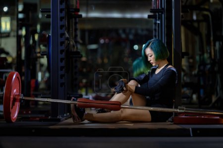 Foto de Mujer asiática se enfrenta al síndrome de sobreentrenamiento después del entrenamiento con pesas dentro del gimnasio con fondo oscuro para el ejercicio y la aptitud - Imagen libre de derechos