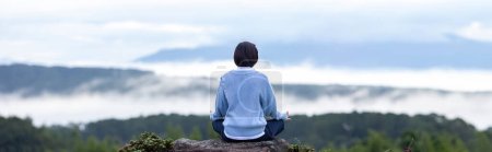 Photo for Panorama back view of woman in hoodie is relaxingly practicing meditation yoga at top of mountain with mist and fog in summer to attain happiness from inner peace wisdom for healthy mind and soul - Royalty Free Image
