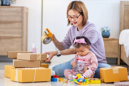 Téléchargez les photos : Jeune mère asiatique emballe paquet de produits de marchandises tout en travaillant à la maison prêt à livrer tandis que son enfant en bas âge joue avec son jouet - en image libre de droit