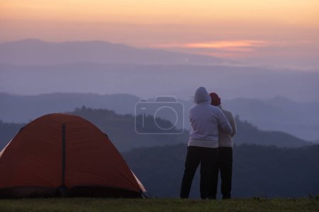 Téléchargez les photos : Couple se serre dans ses bras par la tente pendant la nuit de camping tout en regardant le beau coucher de soleil pittoresque sur la montagne pour des vacances d'aventure en plein air Voyage - en image libre de droit