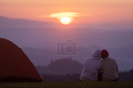 Foto de Pareja está sentada junto a la tienda durante la noche acampando mientras mira la hermosa puesta de sol panorámica sobre la montaña para el concepto de viaje de aventura al aire libre vacaciones - Imagen libre de derechos