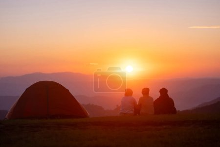 Foto de Grupo de amigos sentados junto a la tienda durante la noche acampando mientras miran el hermoso punto de vista puesta de sol sobre la montaña para el concepto de viaje de aventura al aire libre - Imagen libre de derechos