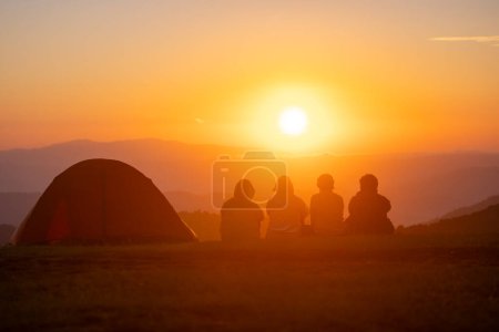 Foto de Grupo de amigos sentados junto a la tienda durante la noche acampando mientras miran el hermoso punto de vista puesta de sol sobre la montaña para viajes de vacaciones de aventura al aire libre - Imagen libre de derechos