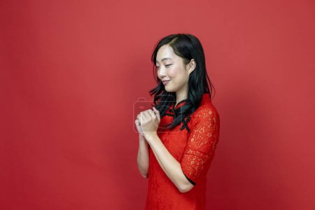 Photo for Asian chinese woman with red cheongsam or qipao doing polite respectful gesture for wishing good luck and prosperity in Chinese New Year celebration holiday isolated on red background - Royalty Free Image