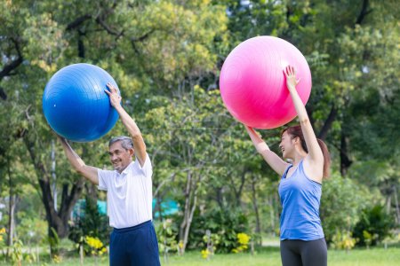 Foto de Hombre asiático mayor y su hija están haciendo ejercicio con bola de yoga en el parque público para construir el núcleo muscular del cuerpo para el ejercicio de longevidad mayor y entrenamiento al aire libre - Imagen libre de derechos