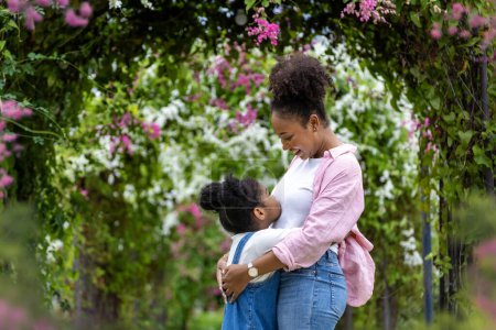 Téléchargez les photos : Mère africaine et sa fille s'embrassant tout en marchant joyeusement sous l'arche en treillis fleuri dans le parc public pendant l'été pour l'amour de la famille et les soins dans la célébration de la fête des mères - en image libre de droit
