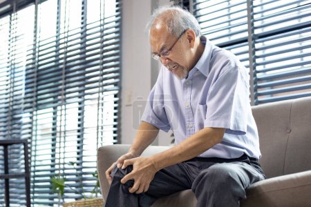 Photo for Senior asian man suffering from knee osteoarthritis symptom whiling sitting on the couch at home for medical surgery treatment and physical therapy - Royalty Free Image