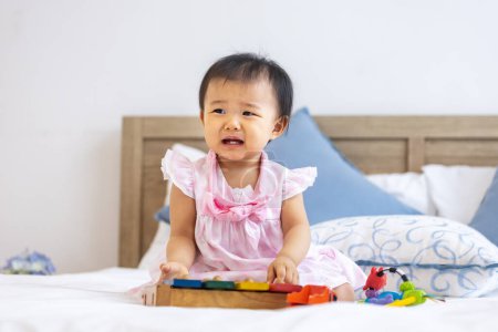 Photo for Adorable little asian baby toddler is sitting on the bed crying after playing with her wooden melody toy for preschool learning and growth development - Royalty Free Image