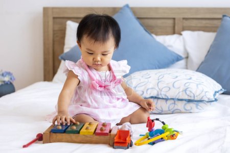 Téléchargez les photos : Adorable petit bébé asiatique est assis sur le lit jouet mélodique en bois pour l'apprentissage préscolaire et le développement de la croissance - en image libre de droit