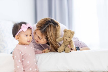 Photo for Asian mother is holding her pretty smiling baby daughter while spending quality time in the bed for family happiness and parenting - Royalty Free Image
