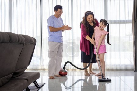 Téléchargez les photos : Famille asiatique avec père, mère et fille s'entraident pour nettoyer la maison à l'aide d'une machine à vide pour les tâches quotidiennes de routine et l'entretien ménager - en image libre de droit