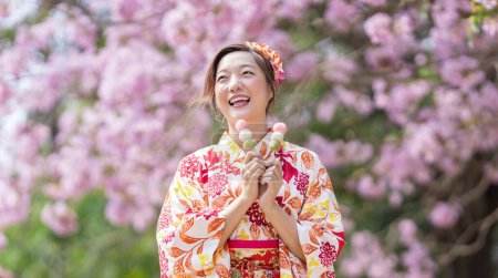 Foto de Mujer japonesa en vestido tradicional de kimono sosteniendo dulce postre de hanami dango mientras camina en el parque en el árbol de flor de cerezo durante el concepto del festival de sakura de primavera - Imagen libre de derechos