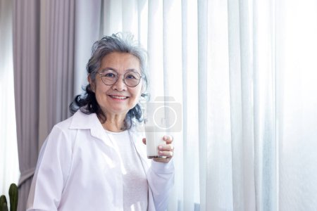 Foto de Señora asiática mayor bebiendo vaso de leche mientras está de pie junto a la ventana con espacio de copia para aumentar el calcio y el consumo de productos lácteos saludables - Imagen libre de derechos