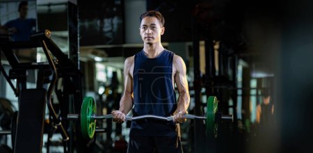 Photo for Asian muscular man is practice weight lifing using easy bar as beginner on barbell for arm and core muscle inside gym with dark background for exercising and workout usage - Royalty Free Image