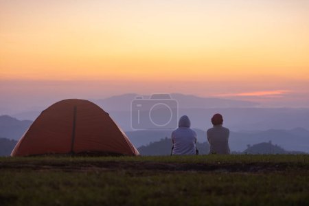 Couple is sitting by the tent during overnight camping while looking at the beautiful scenic sunset over the mountain for outdoor adventure vacation travel