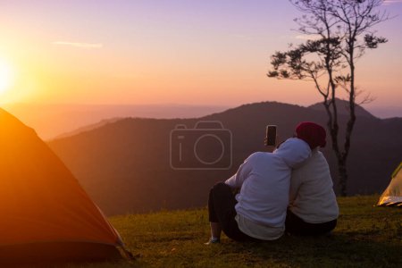 Foto de Pareja está sentada junto a su tienda durante la noche acampando y tomando selfie mientras mira la hermosa puesta de sol panorámica sobre la montaña para viajes de vacaciones de aventura al aire libre - Imagen libre de derechos