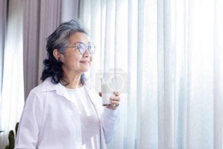 Téléchargez les photos : Dame asiatique senior boit un verre de lait tout en restant près de la fenêtre avec un espace de copie pour stimuler le calcium et une consommation saine de produits laitiers - en image libre de droit