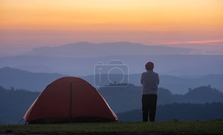 Der Reisende steht während der Zeltübernachtung am Zelt, während er den wunderschönen Sonnenuntergang über dem Berg für eine Abenteuerreise im Freien betrachtet