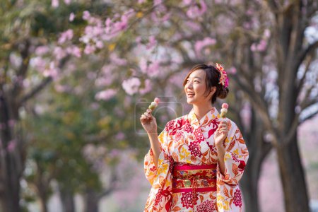 Téléchargez les photos : Femme japonaise en robe de kimono traditionnelle tenant doux hanami dango dessert tout en marchant dans le parc au cerisier pendant le festival de sakura de printemps - en image libre de droit