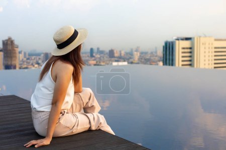 Photo for Pretty tourist woman is relaxing and enjoying her rooftop swimming pool view with cityscape background for vacation and travel usage - Royalty Free Image