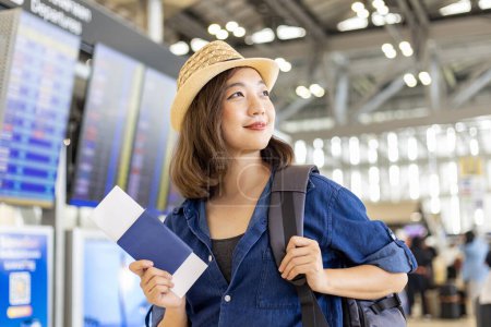 Téléchargez les photos : Jeune femme asiatique passager avec ses bagages à main regarde sa carte d'embarquement après auto-enregistrement kiosque dans le terminal de l'aéroport pour un vol de voyage international et des vacances - en image libre de droit