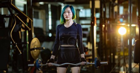 Téléchargez les photos : Femme asiatique est pratique haltérophilie en utilisant barre facile comme débutant sur barre pour bras et noyau musculaire à l'intérieur gymnase avec fond sombre pour l'exercice et le concept d'entraînement - en image libre de droit