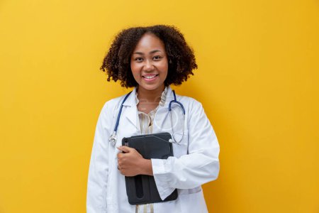 Photo for African American female doctor smiling with full uniform and stethoscope on yellow background for medical and healthcare - Royalty Free Image
