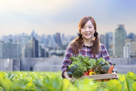 Foto de Mujer asiática jardinero está cosechando verduras orgánicas mientras trabaja en la agricultura urbana en la azotea ciudad futurista jardinería sostenible en el espacio limitado para reducir la huella de carbono y la seguridad alimentaria - Imagen libre de derechos