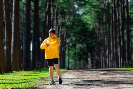 Téléchargez les photos : Chemin asiatique coureur court en plein air dans la forêt de pins route de terre pour l'exercice et l'entraînement des activités tout en vérifiant la montre intelligente sur la perte de calories atteindre un mode de vie sain et de remise en forme - en image libre de droit