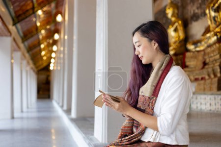 Asian buddhist woman reading Sanskrit ancient palm leaf manuscript of Tripitaka the Lord Buddha dhamma teaching while sitting in temple on holy full moon day to chant and worship inside monastery