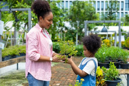 Téléchargez les photos : La mère et la fille africaines choisissent la plante ornementale et les pots de fleurs en fleurs de la pépinière du centre de jardin local pendant l'été pour le jardinage de week-end et le plein air - en image libre de droit