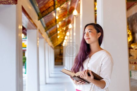 Asian buddhist woman is reading Sanskrit ancient Tripitaka book of Lord Buddha dhamma teaching while visiting temple for chanting and worship inside the monastery