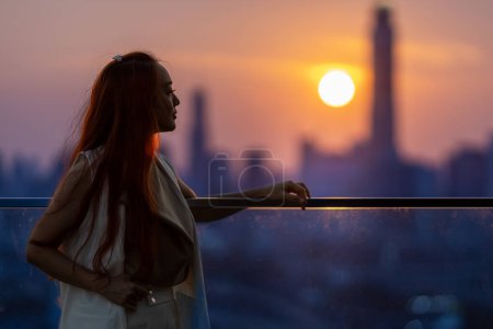Frau schaut und genießt den Sonnenuntergang vom Balkon mit Sonnenuntergang hinter Wolkenkratzer in der geschäftigen Innenstadt mit Einsamkeit für Einsamkeit, Einsamkeit und Traum von Freiheit Lebensstil