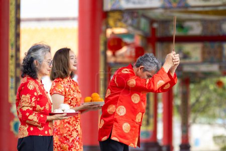 Asiatische Familie mit älteren Eltern in rotem Cheongsam-Qipao-Kleid bietet dem Ahnengott im chinesischen buddhistischen Tempel während des Mondneujahres Essen an und wünscht Segen und viel Glück