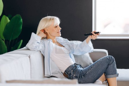 Happy young blonde woman switches channels on TV trying to choose something interesting.