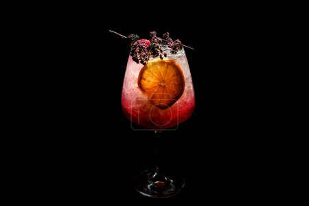 Foto de Copa de cóctel rojo helado servido en una copa de vino, decorado con rodajas de naranja y rama de romero. Fondo negro. - Imagen libre de derechos