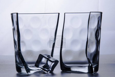 Foto de Vidrio roto en dos mitades con un cubo de hielo. Fotografiado sobre fondo blanco en estudio. Bodegón. - Imagen libre de derechos