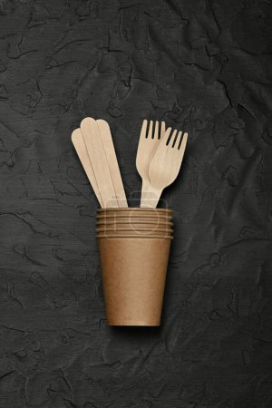 Photo for Close up disposable brown paper takeaway coffee cup, wooden spoons and forks on black background, natural eating and drinking utensils, close up, elevated table top view, directly above - Royalty Free Image