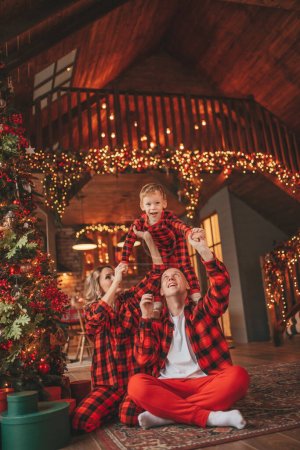 Smile active parents with small son in red checkered sleepwears waiting Santa indoor. Stylish family love laughing hugs kisses at home. Celebrate New year garlands and lights eve 25 December Noel tree
