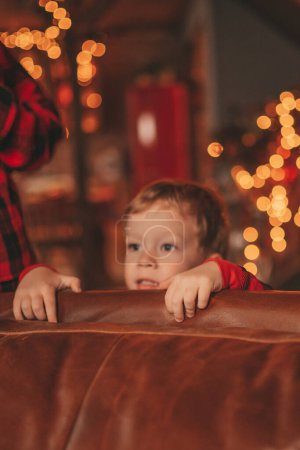 Photo for Smile small child being active having fun in motion and waiting for miracle Santa. Red checkered sleepwear kid playing indulge indoor jumping moving laughing garlands lights Noel tree eve 25 December - Royalty Free Image