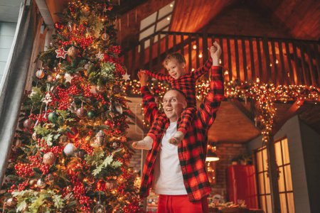 Photo for Smile little boy sits on father neck having fun with each other indoor waiting miracle. Celebrating New year with garlands lights of Noel tree winter season Xmas spirit eve 25 December paternal love - Royalty Free Image