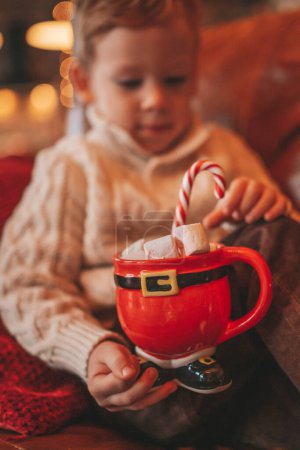 Photo for Happy little boy dreamer waiting at miracle Santa enjoy sweets color lollipop. Cheerful kid in knitwear outfit celebrating new year hold Xmas cup with marshmallows at bokeh lights noel eve 25 december - Royalty Free Image