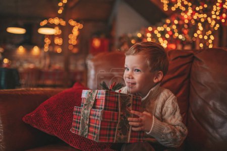 Photo for Smile small child being active having fun waiting for miracle Santa open presents. Cheerful kid in casual knit outfit celebrating new year with gifts boxes garlands bokeh lights noel eve 25 december - Royalty Free Image